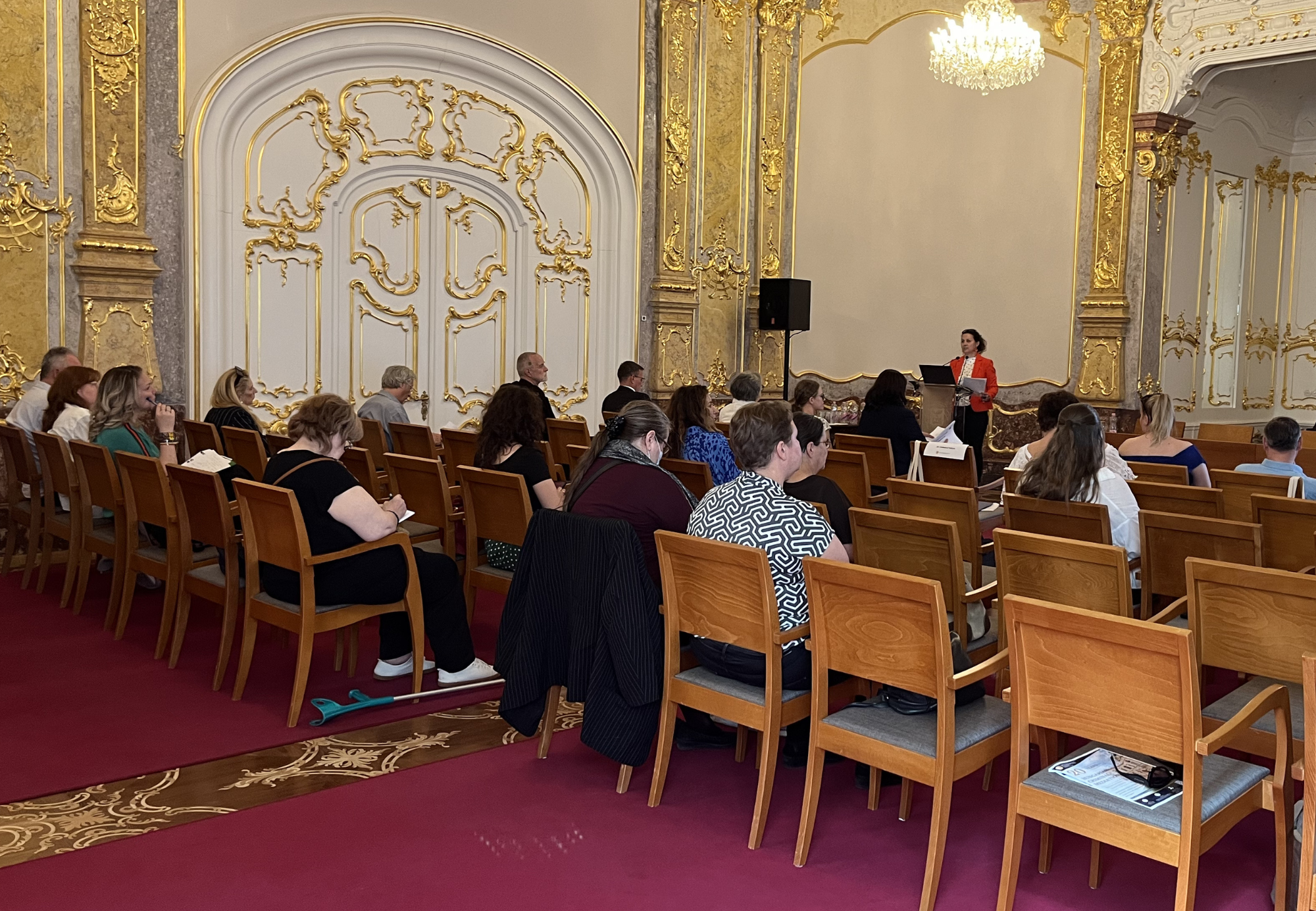 A képen A képen Dr. Dobrocsi Szilvia előadást tart a 20 éves a magyar büntetőjogi mediáció című nemzetközi konferencián. Az előtérben a hallgatóság látható hátulnézetből, amint figyelemmel kíséri az előadást.